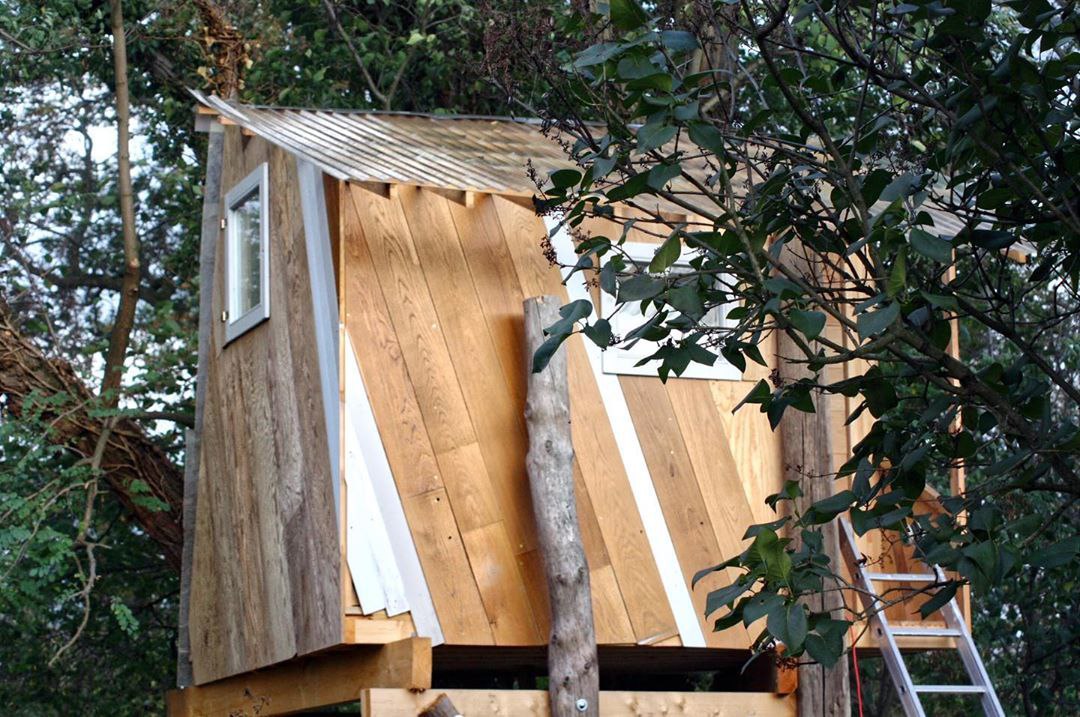 Boomhut van eikenhouten planken met spits dak tussen de bomen
