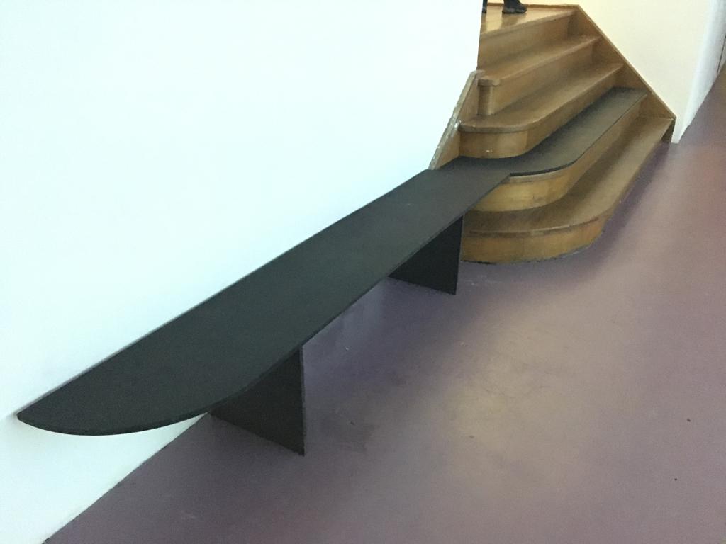 Zwarte stalen gebogen bank naast een houten trap in een tentoonstellingsruimte